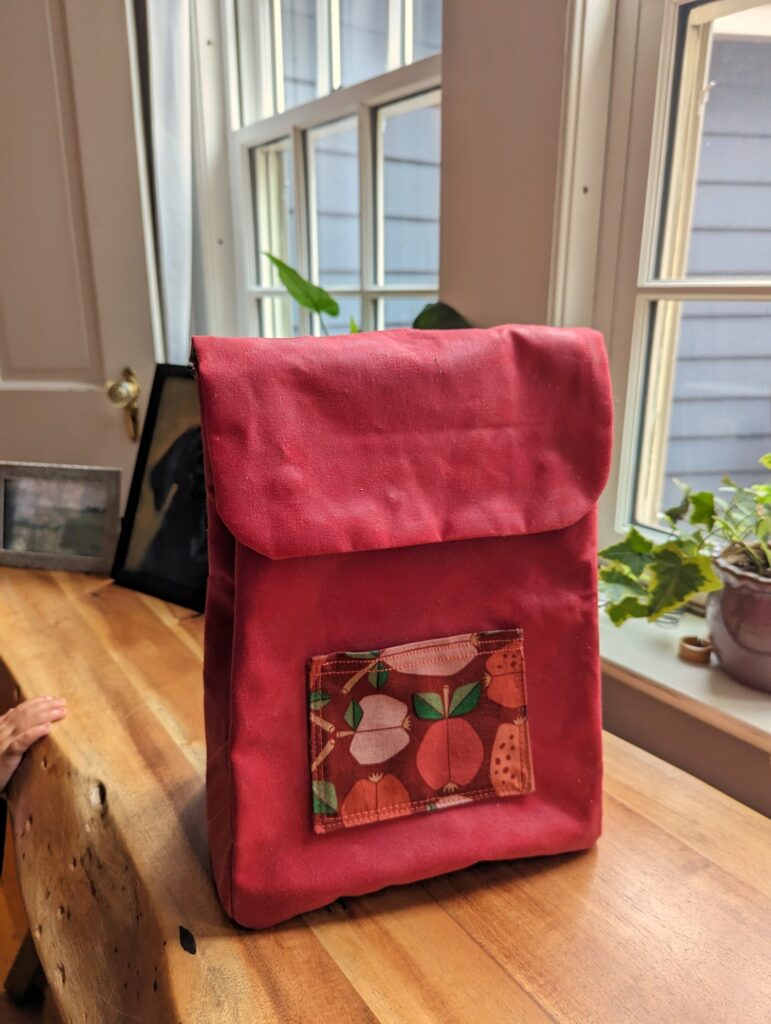 Red toddler backpack viewed from the front. It has a small patch pocket with apple patterned fabric.
