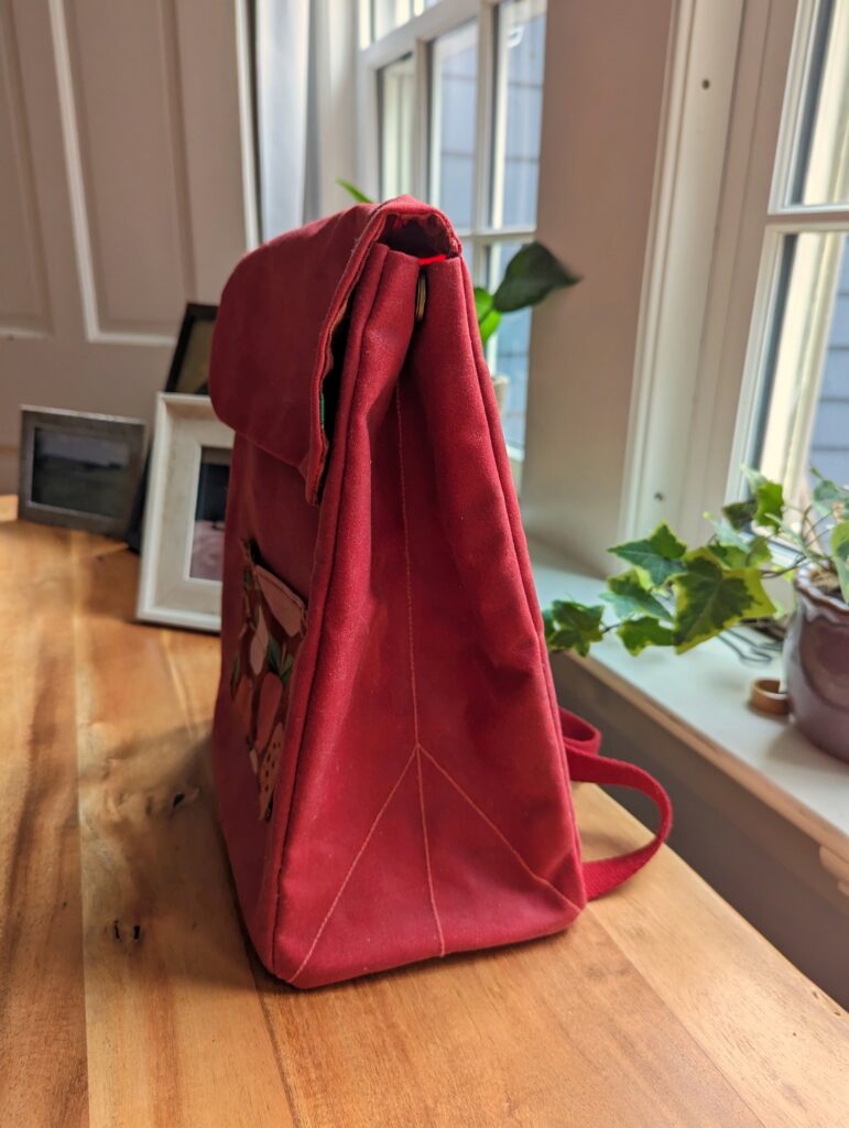Red toddler backpack viewed from the side. The side folds in like a paper bag, and the top flap folds over and snaps onto the front.