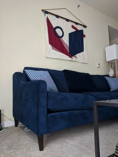 Photo of a living room. There is a blue couch against a cream-colored wall. On the couch are pillows with a blue and red triangle pattern. On the wall above the couch is a fabric wall hanging with an abstract design of geometric shapes in blue and red.