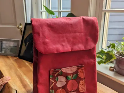 Red toddler backpack viewed from the front. It has a small patch pocket with apple patterned fabric.