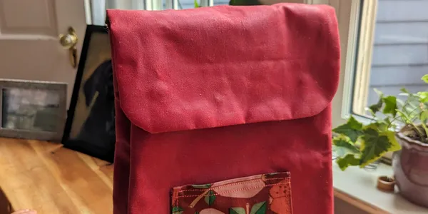 Red toddler backpack viewed from the front. It has a small patch pocket with apple patterned fabric.
