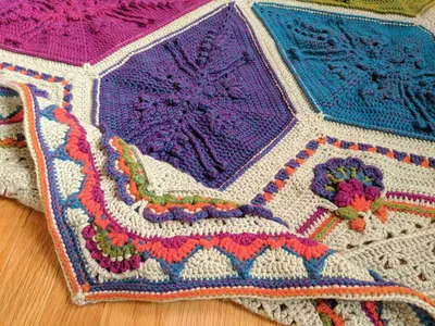 A top-down image of an Ubuntu blanket laid flat on a wood floor. The blanket is hexagonal. At the center, there are 6 large diamonds arranged in a star shape. Each diamond is a different bright color (orange, green, blue, purple, magenta, salmon). To fill out the sides and make a hexagon, there are triangle shapes in between each pair of diamonds. These side triangles have elaborate flowers on them made of the same colors as the diamonds on a light gray background. All the way around the hexagon is an elaborate colorful border.