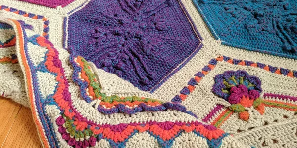 A top-down image of an Ubuntu blanket laid flat on a wood floor. The blanket is hexagonal. At the center, there are 6 large diamonds arranged in a star shape. Each diamond is a different bright color (orange, green, blue, purple, magenta, salmon). To fill out the sides and make a hexagon, there are triangle shapes in between each pair of diamonds. These side triangles have elaborate flowers on them made of the same colors as the diamonds on a light gray background. All the way around the hexagon is an elaborate colorful border.