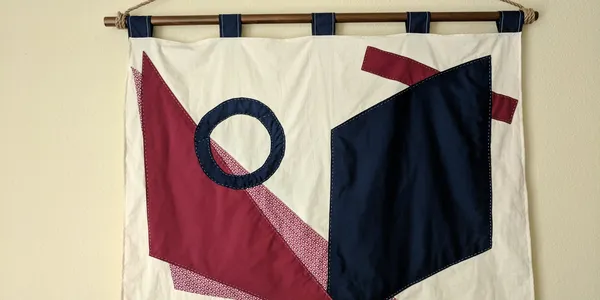 Photo of a living room. There is a blue couch against a cream-colored wall. On the couch are pillows with a blue and red triangle pattern. On the wall above the couch is a fabric wall hanging with an abstract design of geometric shapes in blue and red.