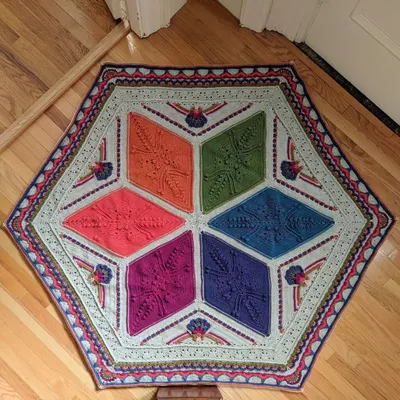A top-down image of an Ubuntu blanket laid flat on a wood floor. The blanket is hexagonal. At the center, there are 6 large diamonds arranged in a star shape. Each diamond is a different bright color (orange, green, blue, purple, magenta, salmon). To fill out the sides and make a hexagon, there are triangle shapes in between each pair of diamonds. These side triangles have elaborate flowers on them made of the same colors as the diamonds on a light gray background. All the way around the hexagon is an elaborate colorful border.