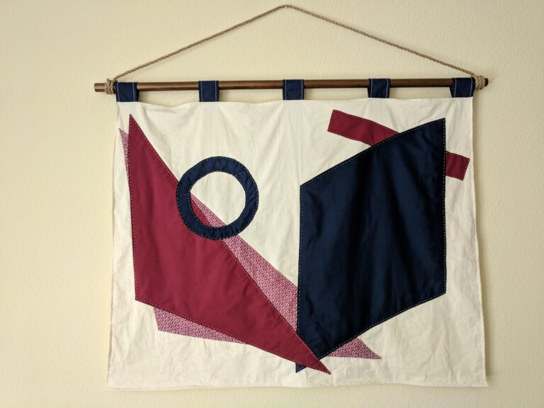 Photo of a fabric wall hanging. The background is a rectangular piece of off-white fabric, upon which are layered geometric shapes in navy blue and dark red. The shapes are sewed to the background with an intentionally visible white running stitch. It is hanging from a wooden post with large blue tabs.