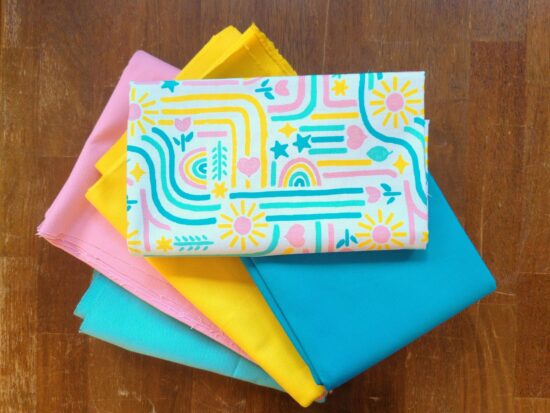 Canvas fabric with bright pink, yellow, blue, and green hand-drawn patterns folded up and stacked on matching solid color canvas.